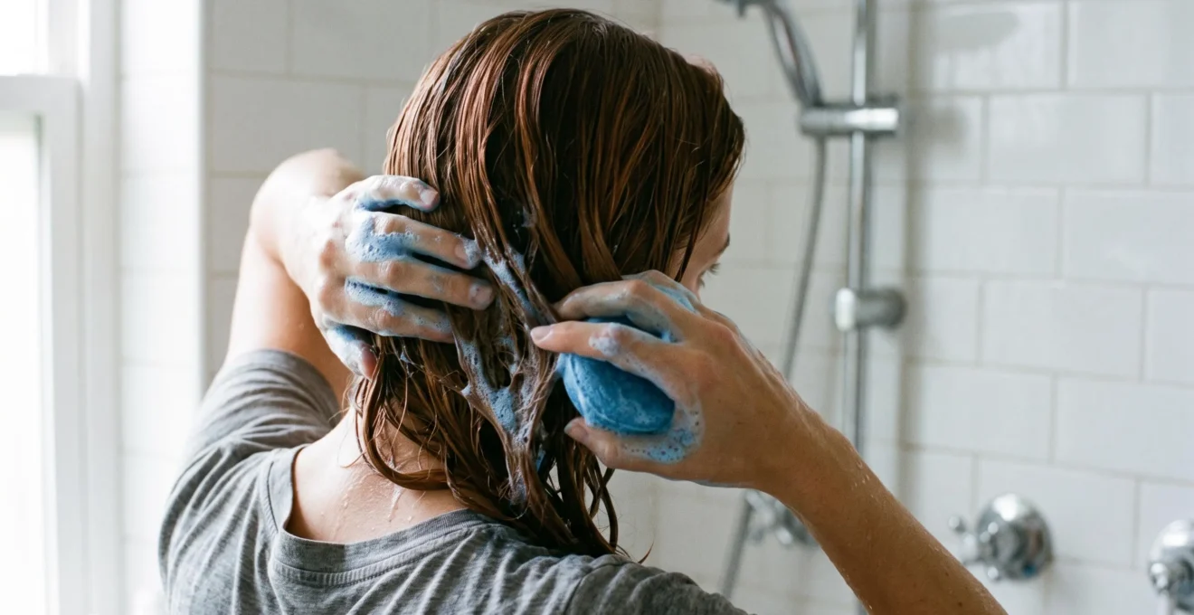 Personne aux cheveux châtains appliquant un shampoing solide bleu avec mousse bleue visible dans les longueurs mouillées sous la douche